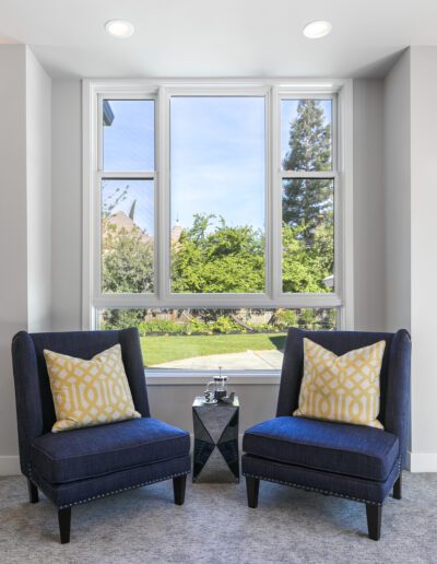 Two navy blue chairs with yellow patterned pillows face a window overlooking a green garden, with a small black side table between them.