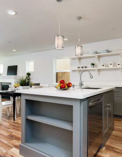 Modern kitchen with a central island, white countertops, gray cabinets, and wood flooring. Dining area and living room with a couch visible in the background.