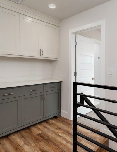 Modern hallway with wooden floors, light gray upper cabinets, dark gray lower cabinets, and black metal railing. A white door is partially open in the background.