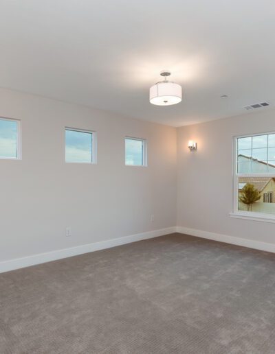 Empty room with beige walls, carpeted floor, three small windows, and one large window. Ceiling and wall lights are present. A view of houses is visible through the large window.