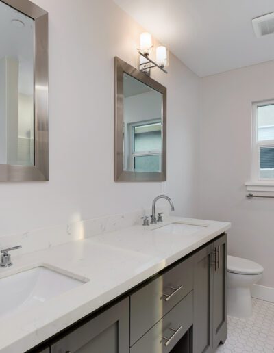 Modern bathroom with double sinks, rectangular mirrors, and a marble countertop. There's a toilet and a window in the background. White tile flooring and walls create a clean, bright look.
