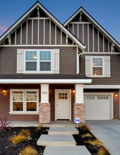 A two-story modern house with brown and white exterior, front porch, and landscaped yard. The driveway leads to a single garage door.