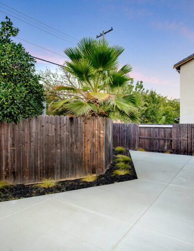 A fenced backyard with concrete patio, wooden fence, and small plants lining the edge.