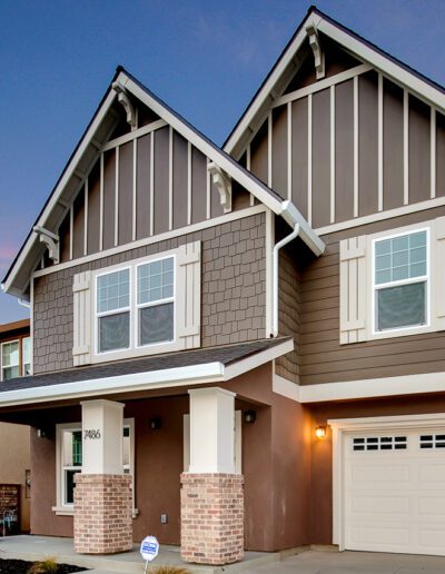 Two-story house with a modern design, featuring brown and gray siding, white trim, and a single-car garage. The house is illuminated with outdoor lighting under a twilight sky.