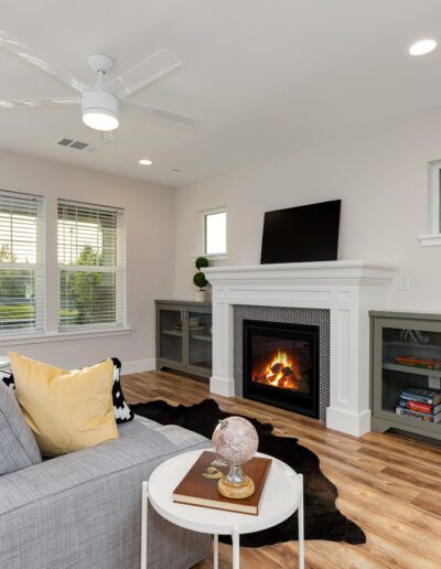 Modern living room with a gray couch, yellow pillow, white ceiling fan, and a fireplace. Two cabinets with books and decor flank the fireplace. Large windows provide natural light.