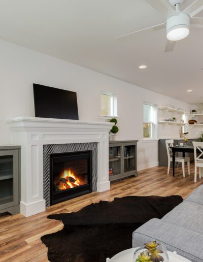 Modern living room with a fireplace, mounted TV, gray cabinets, and a sofa. Open kitchen and dining area with barstools and a ceiling fan. Dark rug on wooden floor.