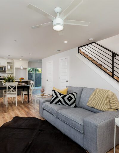 Open-concept living area with a gray couch, black and white pillows, wooden flooring, staircase with a black railing, and a dining area near a modern kitchen in the background.