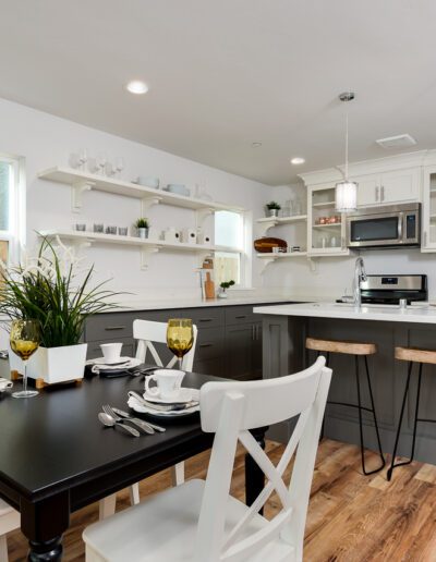 Modern kitchen and dining area with white cabinets, black dining table set for four, stools at a kitchen island, wooden floor, and pendant lights.